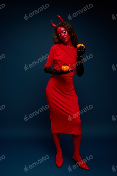 Preview: A stunning woman in a vibrant Halloween costume posing with pumpkins in a dark setting