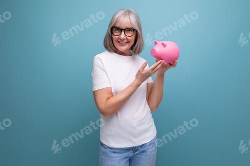 Preview: grey-haired grandmother keeps her pension in a piggy bank on a studio background with copy space