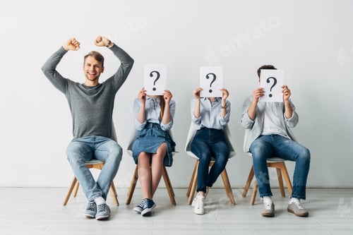 Preview: Cheerful man showing yeah gesture near candidates with question marks on cards in office