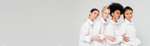 Preview: young multiethnic women wearing white shirts and looking at camera isolated on grey,banner
