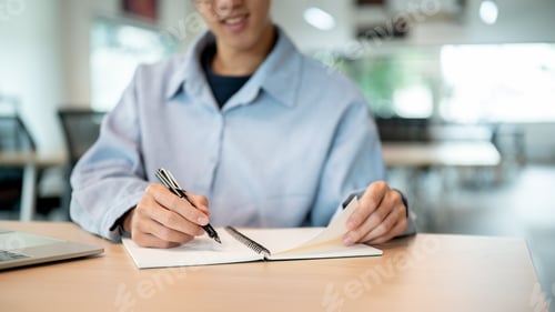 Preview: Smiling asian man holding a fountain pen writing in notebook while sitting at wooden table in cafe.