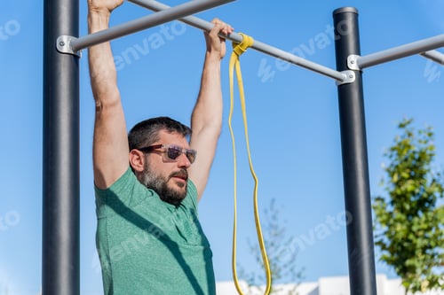 Preview: man with beard and sunglasses training his back by doing pull-ups on a bar in an outdoor gym