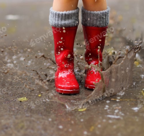 Preview: Child wearing red rain boots jumping into a puddle. Close up