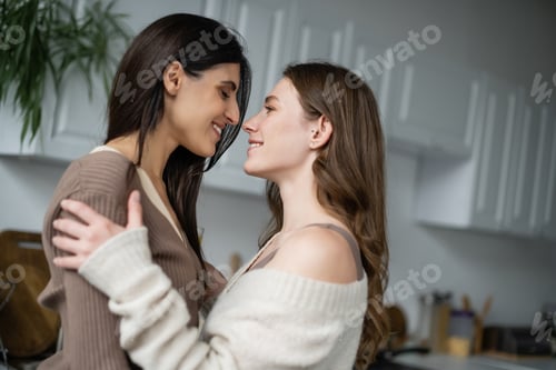 Preview: Side view of cheerful lesbian women hugging in blurred kitchen