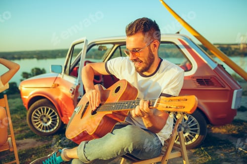 Preview: Couple sitting and resting on the beach playing guitar on a summer day near river