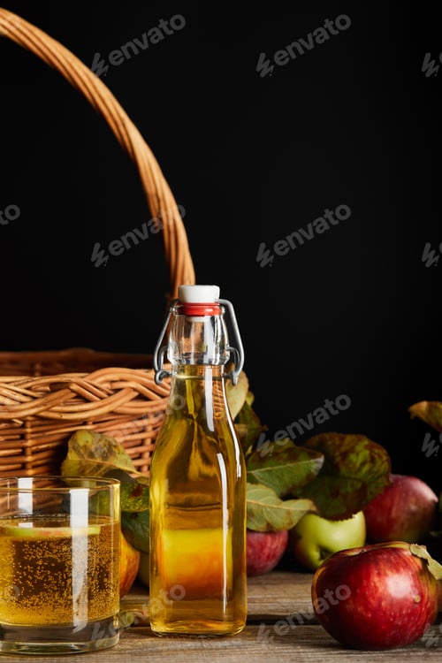 Preview: bottle and glass of cider near wicker basket and apples on wooden surface isolated on black