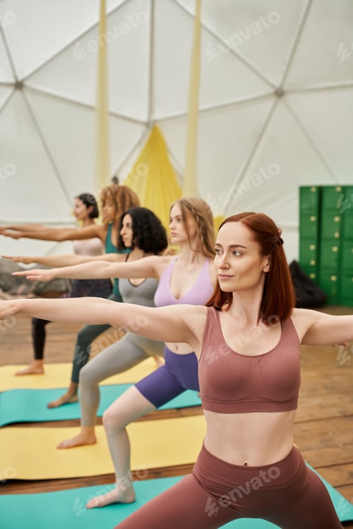 Preview: woman looking away while practicing yoga in warrior pose near multiethnic girlfriends