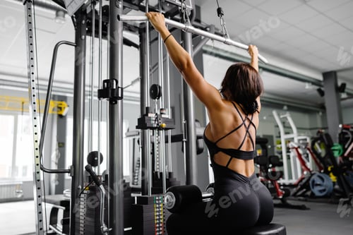 Preview: Muscular athletic female bodybuilder in black suit pulled on sports simulator in gym.