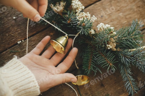 Preview: Making modern christmas wreath. Hands decorating xmas boho wreath with bells on rustic table