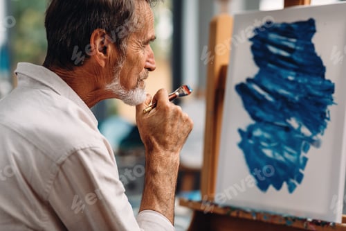 Preview: Close up of a pensive mature painter holds a paintbrush looking at a picture on the easel