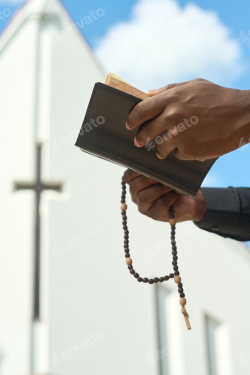 Preview: Hands of black man holding open Holy Bible in leather cover and rosary beads