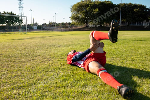 Preview: Male soccer player lying on grass holding knee beside goal frame on outdoor pitch