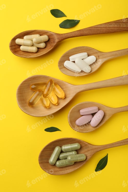 Preview: Different vitamin pills in spoons and green leaves on yellow background, flat lay