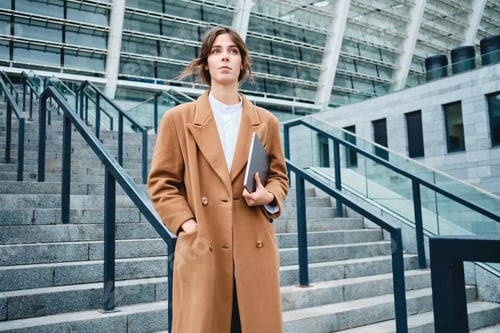 Preview: Young attractive stylish businesswoman in coat with laptop intently looking away outdoor