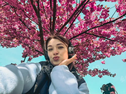 Preview: pretty smiling woman listening music in headset taking selfie under blooming sakura tree