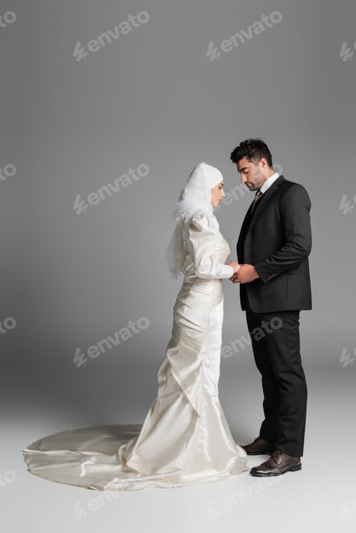 Preview: full length of muslim bride in wedding dress and groom in suit holding hands on grey