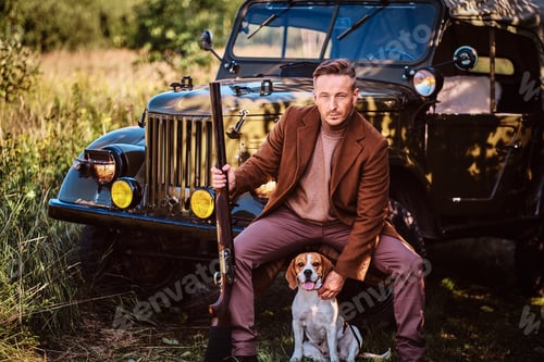 Preview: Hunter in elegant clothes together with his beagle dog next to a retro military car in the forest.