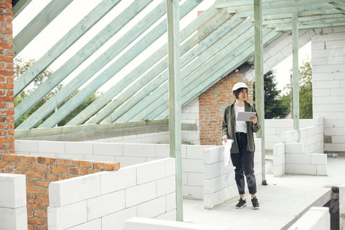 Preview: Young female architect with tablet checking blueprints at wooden roof framing of modern farmhouse