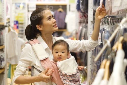 Preview: Asian mother holding her baby in hip seat is choosing the clothes in garment store shop.