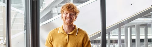 Preview: cheerful ginger man smiling sincerely looking at camera with window on background, coworking, banner