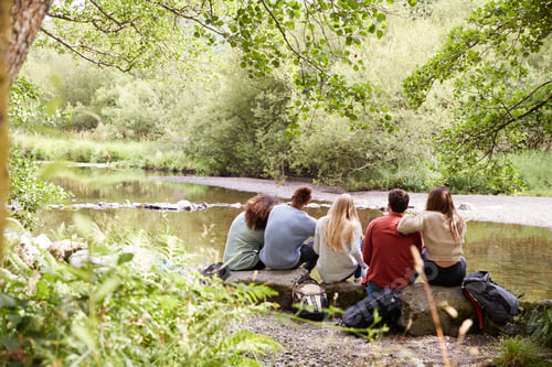 Preview: Five young adult friends taking a break sitting on rocks by a stream during a hike, back view