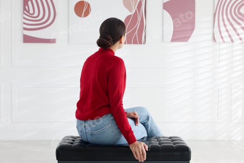 Preview: Woman sitting on bench and looking at paintings in art gallery, back view