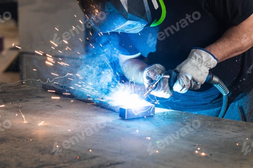 Preview: Welder in protective helmet welding metal detail in workshop