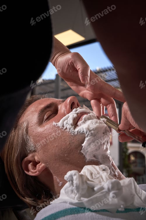 Preview: Crop barber shaving beard of adult client with razor