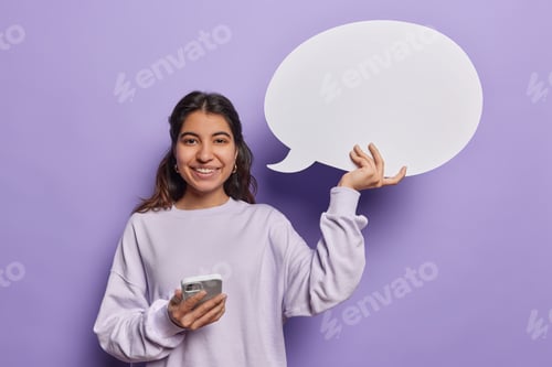 Preview: Waist up shot of pretty cheerful Iranian woman uses modern smartphone holds empty speech bubble