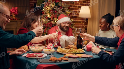 Preview: Joyful man disguised as Santa Claus clinking champagne glass with family enjoying Christmas dinner
