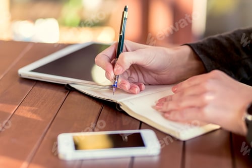 Preview: Person writing in a notebook while using a tablet and smartphone on a wooden table during daylight