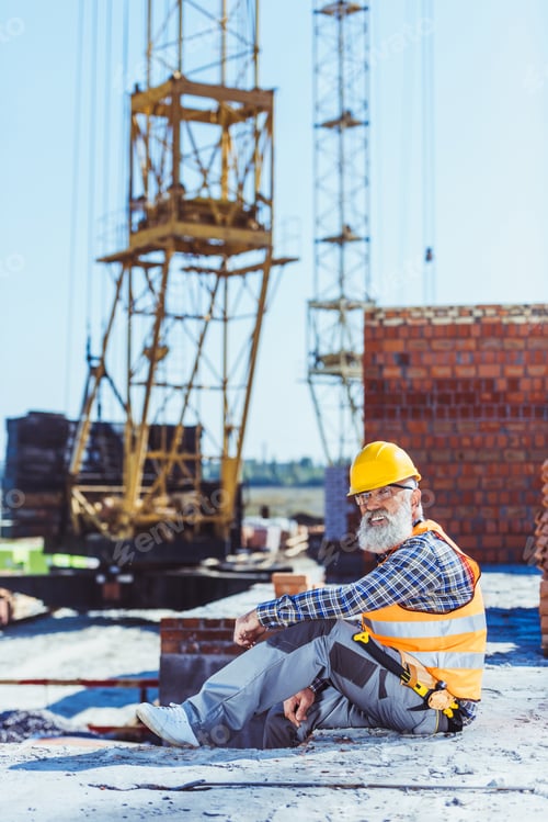 Preview: Bearded worker in reflective vest and hardhat sitting at construction site and looking at camera