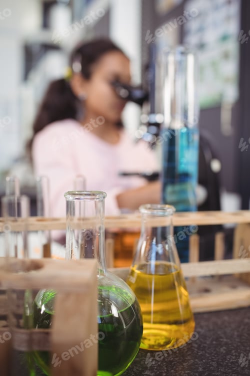 Preview: Flasks of chemicals on desk with elementary student using microscope in background