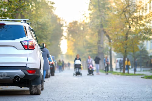 Preview: Car parked on a city street side on bright autumn day with blurred people walking on pedestrian zone