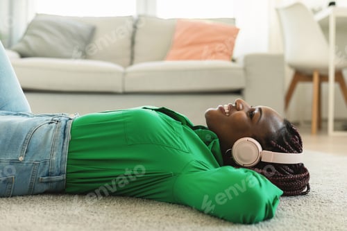 Preview: Young relaxed black lady in wireless headphones listening to music or audio book, lying on floor at