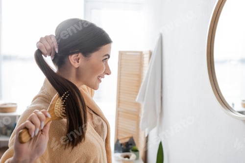 Preview: side view of happy young woman brushing shiny hair and looking at mirror in bathroom