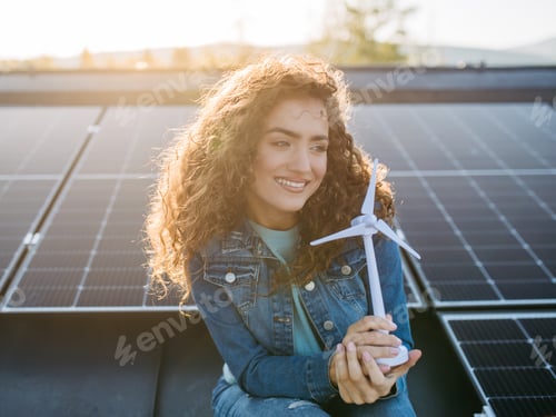 Preview: Portrait of young woman holding model of wind turbine on roof with solar panels.