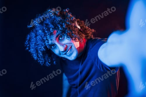 Preview: Takes selfie. Young beautiful man with curly hair is indoors in the studio with neon lighting