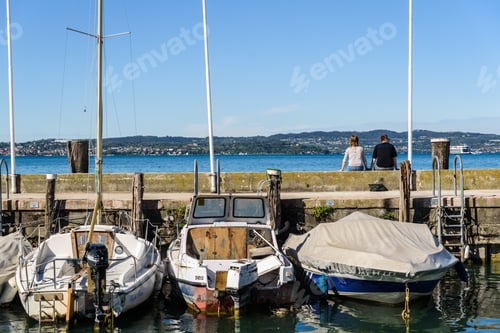 Preview: A couple rest in the port of Sirmione among the hulls of sailboats.