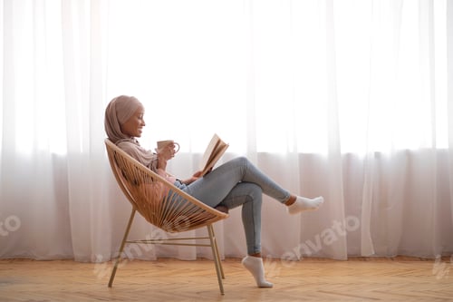 Preview: Side view of young black woman in hijab drinking coffee and reading in cozy armchair indoors, copy