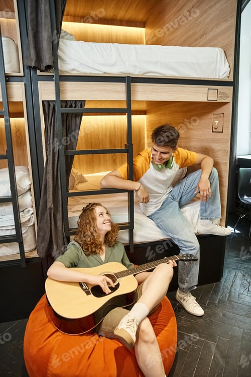 Preview: smiling woman playing acoustic guitar to boyfriend sitting on double-decker bed in hostel room