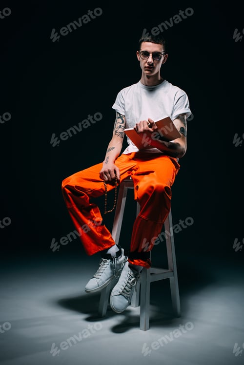 Preview: Serious man in orange uniform with cuffs on legs reading bible and sitting on stool on dark