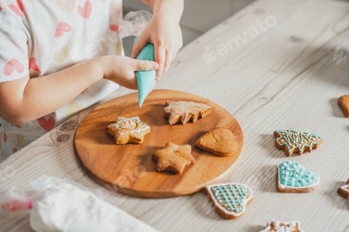 Preview: Partially blurred hands of little girl decorate gingerbread cookies with blue icing in kitchen