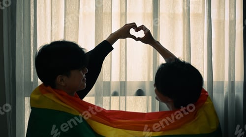 Preview: Romantic young gay couple making heart with their hands while sitting under rainbow flag.