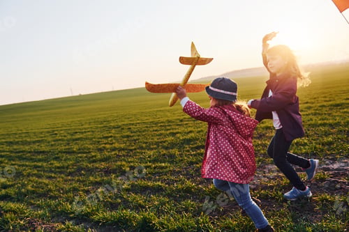 Preview: Two little girls friends have fun together with kite and toy plane on the field at sunny daytime