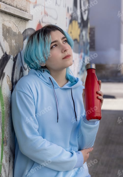 Preview: Blue haired Teenage girl in blue hoodie staying near graffiti wall with red water bottle