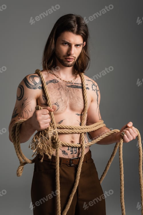 Preview: tattooed and shirtless young man holding rope isolated on grey
