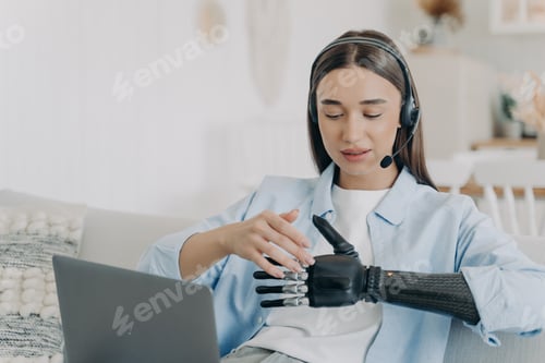 Preview: Handicapped young woman with artificial arm in headset at home. Freelancer has online meeting.