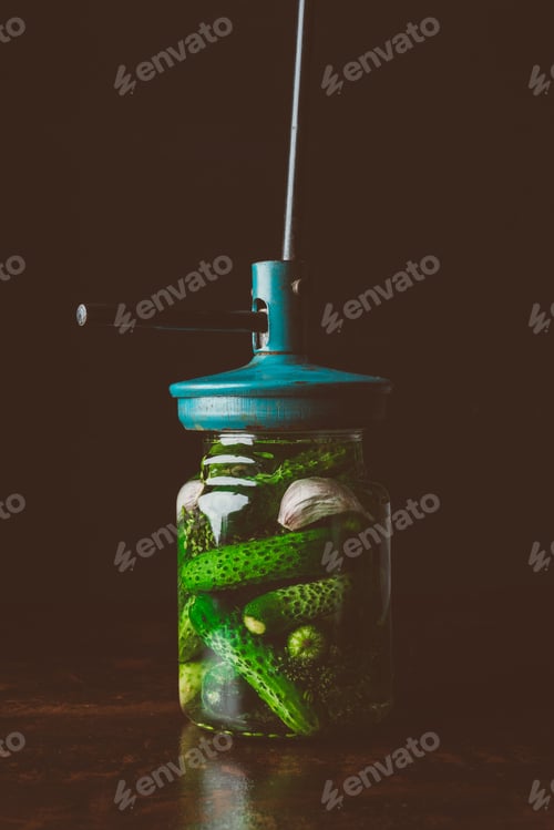 Preview: preserved cucumbers and tool on top of jar on wooden table