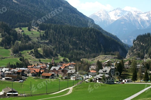 Preview: View of an alpine village nestled in green valleys with snow-capped mountains in the background.